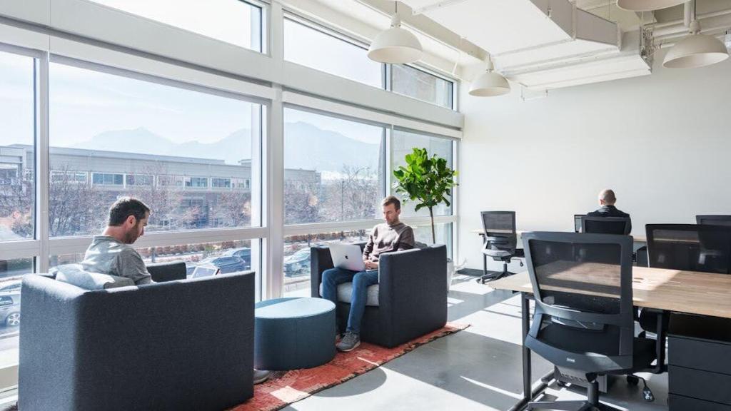 Two people sit in armchairs near a window while another person sits in an office chair at a long table with a potted plant nearby; outside the window is a building and mountains are on the horizon behind the building.