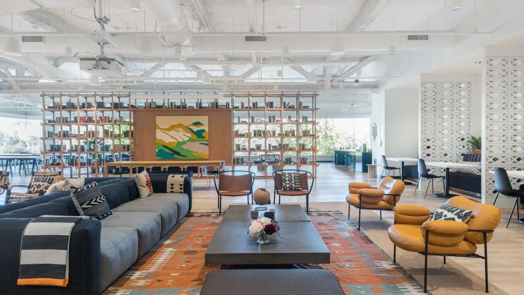 A large office space painted mostly white, with a large bookshelf with several books in the middle of the room and artwork on one side; couches and armchairs with coffee tables are in the foreground with potted plants and cut flowers in some areas; windows line the far end of the room.