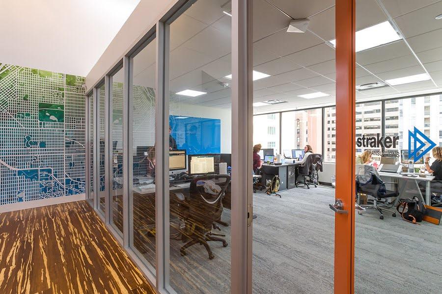 A hallway with woodgrain patterned flooring and a large street map on one wall; a glass wall and orange door lead into an open office.