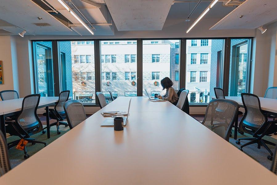 Three long conference tables with a person working on a laptop at the far end of the center table and a tag still attached to a chair.