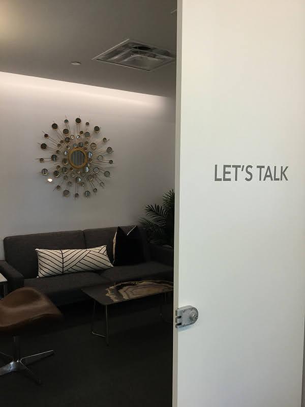 Sun-shaped art with circular mirrors on wall above a couch with coffee table, potted plant, and office chair; a white door reads let's talk.