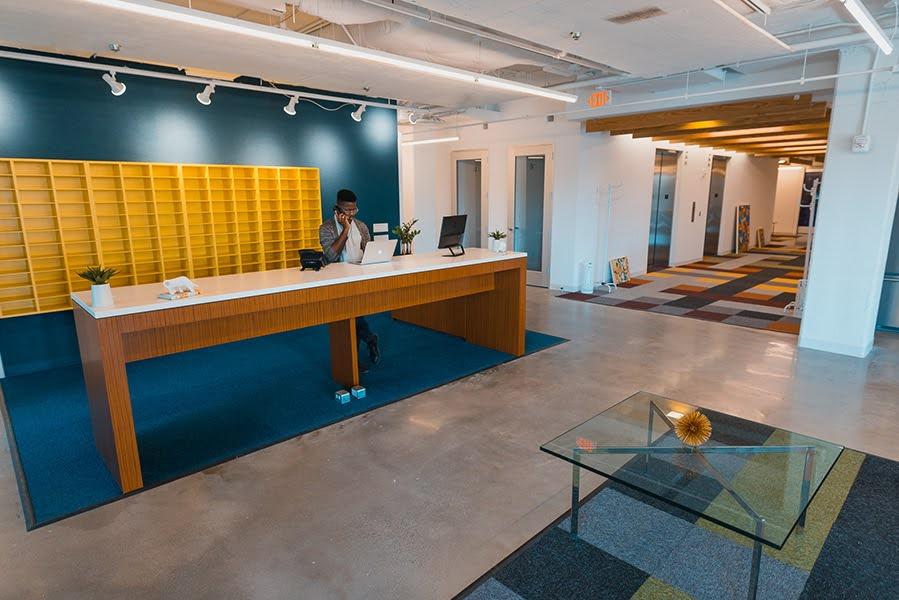 A long reception desk with blue carpeting and yellow shelving along a wall with a person on the phone looking down at a laptop.