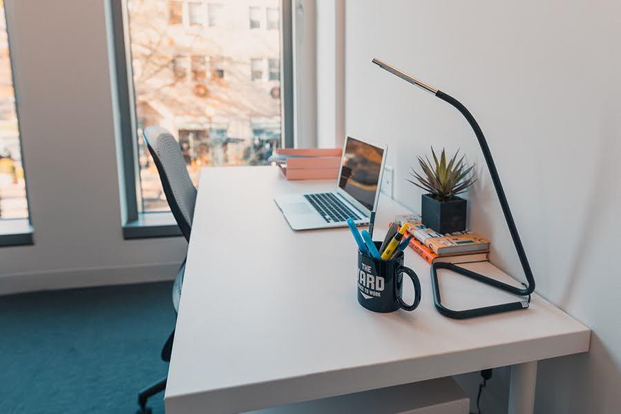 A white desk with an office chair and laptop, potted plant, narrow lamp, books, and a mug containing pens and markers in front of a window.