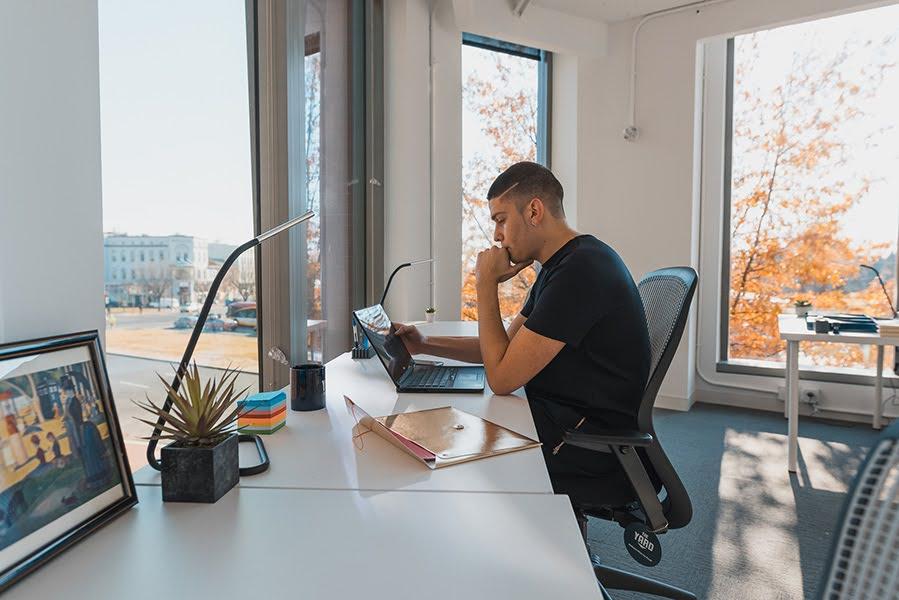 A person sits at a desk with their hand at their chin looking at a laptop screen, with a framed picture in the foreground, lamps and plant.