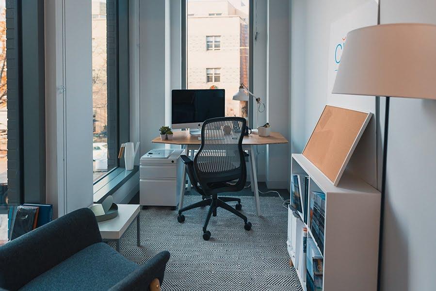 A narrow corner office with diamond pattern carpet, a bookshelf along one wall with a cork board on top, an upholstered armchair.