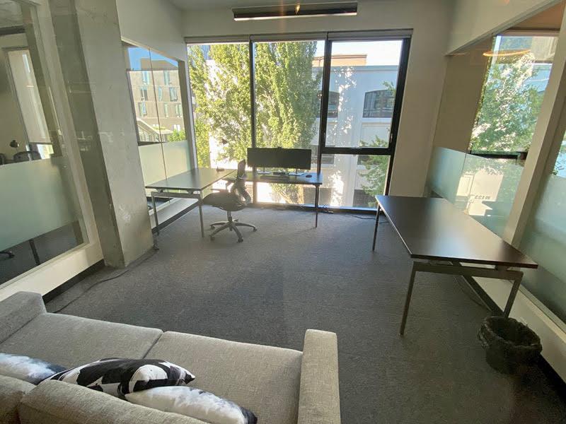 A couch in the foreground with pillows on it and an L-shaped desk with computer monitors along a window at the far end of a working area.