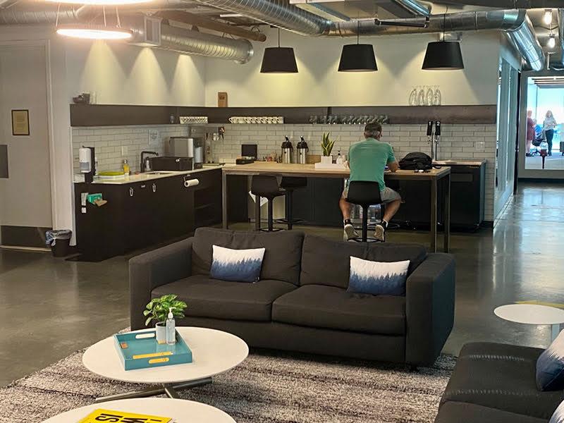 A room with concrete floors and a kitchen area with subway tile; shelves hold mugs and glasses, and there are two taps at one end of a bar. 