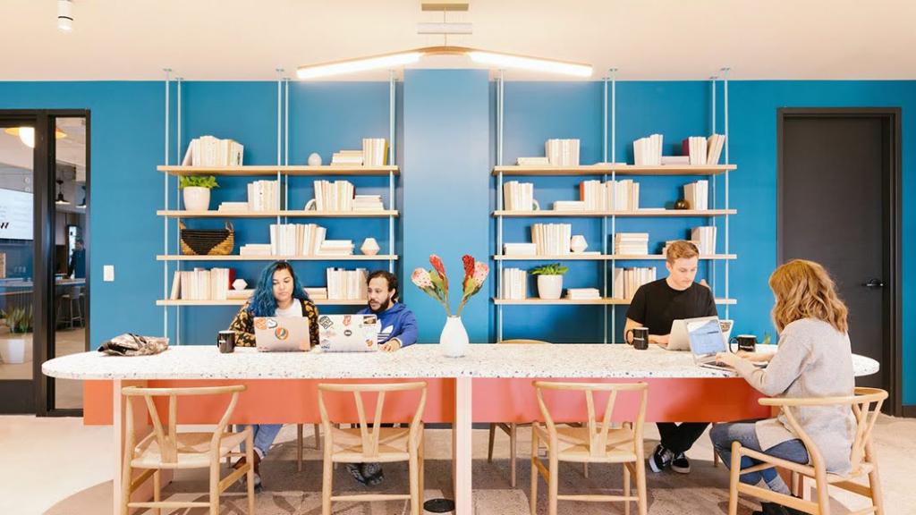 A symmetrical, colorful working space with a long, speckled conference table with wood framed chairs, a flower arrangement