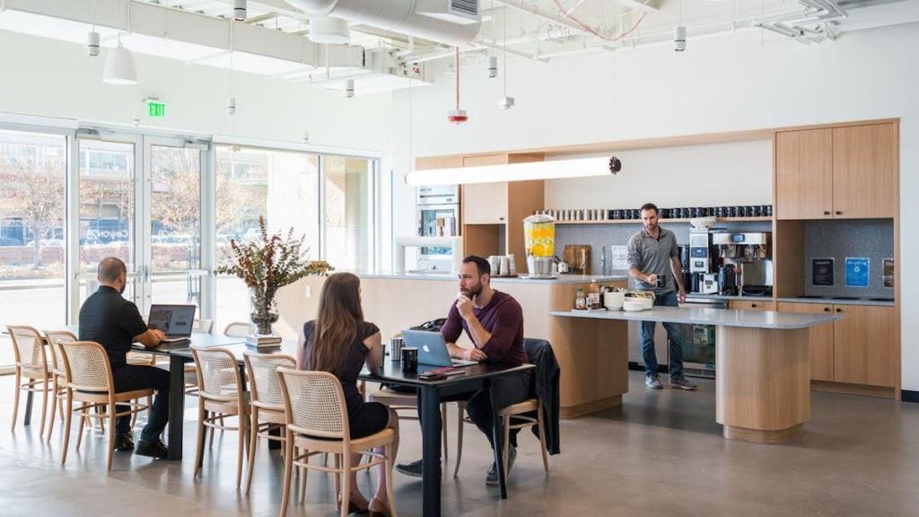 A kitchen area with multiple coffee mugs and cups, cabinets, and a bar; people sit with notebooks and laptops at a long table with a plant.