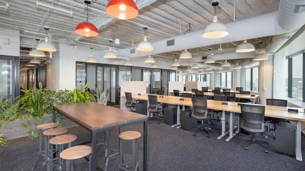 A large office space with windows on one side and long tables with several workstations at each; a hallway at one side of the room.