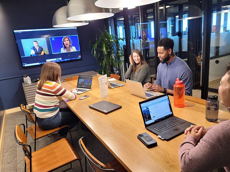 A conference room with dark blue walls and a large flatscreen at one end with two people on screen; four people are gathered around a table.