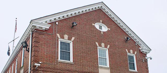 Close up of red brick building facade with three windows and an oval vent above the center window, with U.S. flag in back at left